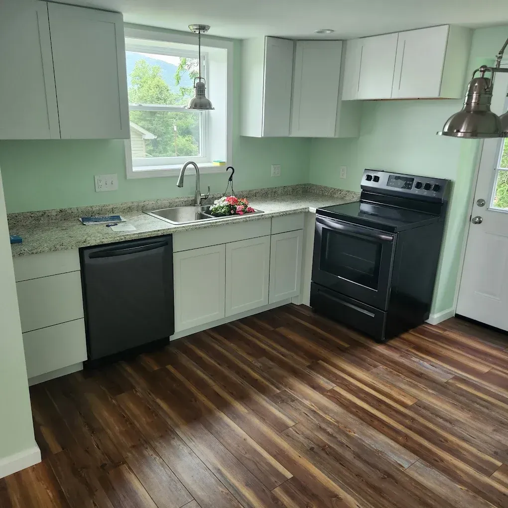 Bright kitchen with white cabinets, black appliances, green walls, and wood flooring.