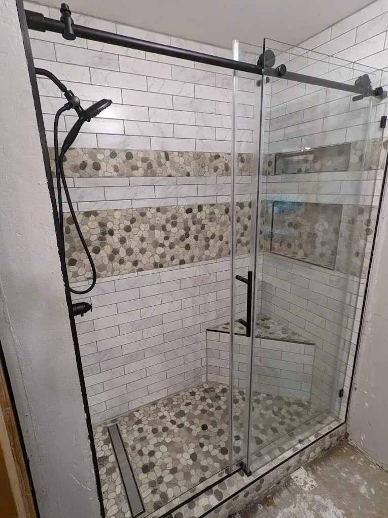Glass shower stall with chrome frame, mosaic tile accents, and a black handheld showerhead in a tiled bathroom