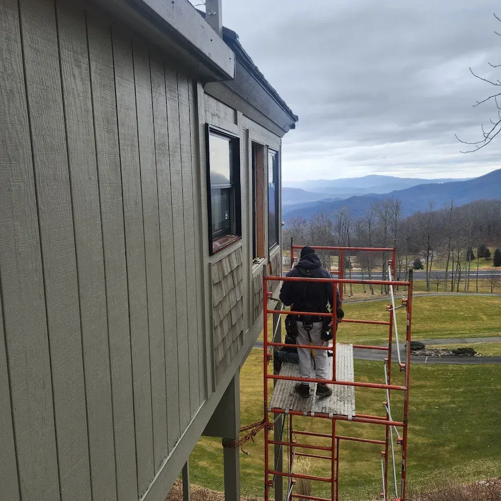 Worker on red scaffolding painting the side of a gray house with mountains in the background