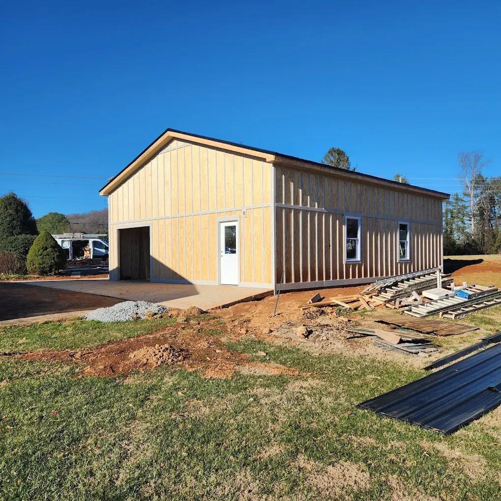 Partially finished tan metal building with garage door, windows, and construction materials on a dirt lot