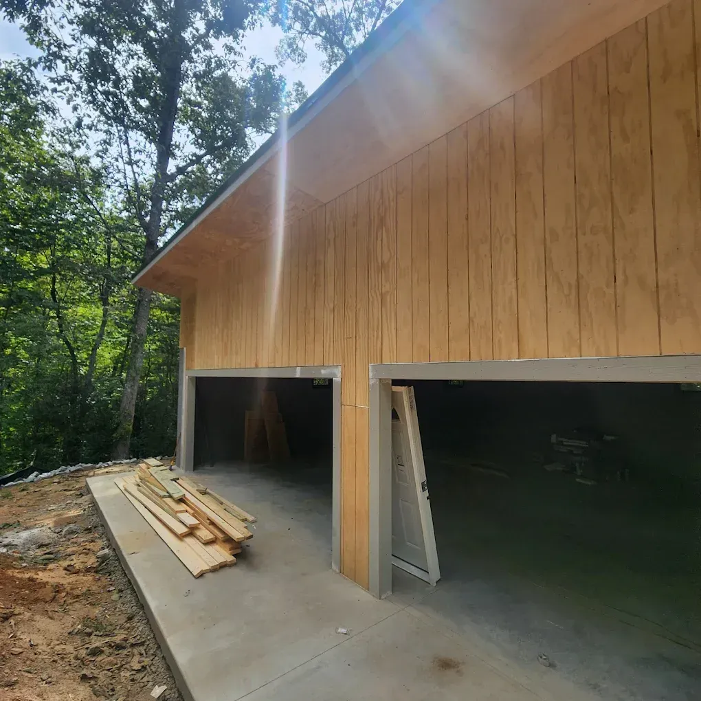 Wood-sided garage under construction with two open bays, lumber stacked outside, and trees surrounding it.