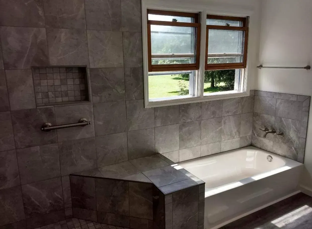 Tiled bathroom with a white bathtub beneath two windows and a built-in shelf in the shower area