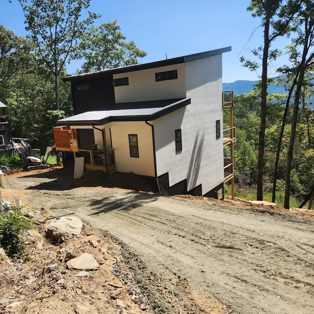 White two-story house on a steep dirt road in a wooded hillside setting