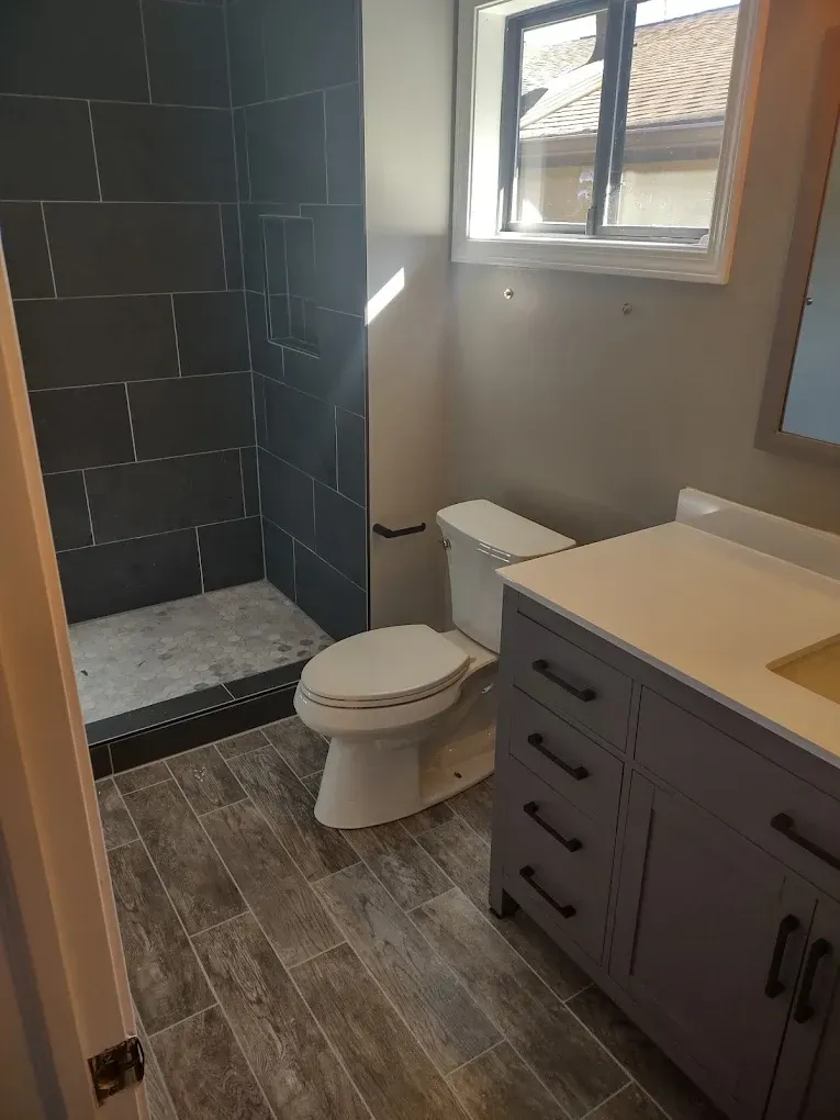 Small bathroom with dark tiled shower, toilet, sink vanity, and window light.