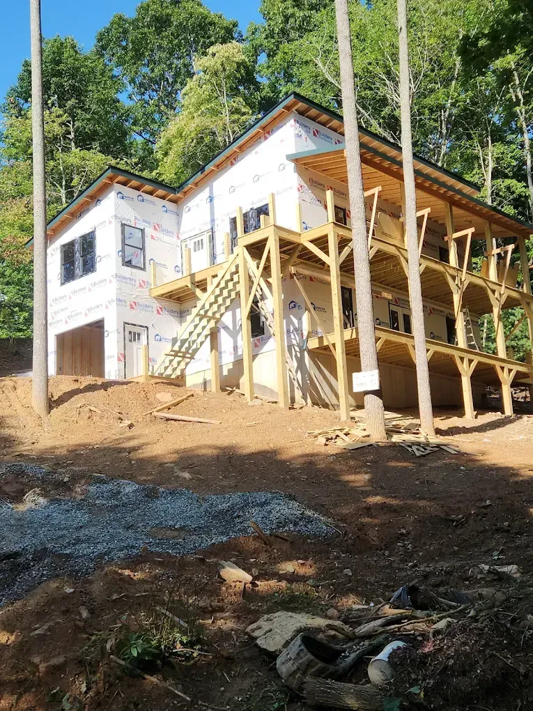 White house under construction on a steep dirt hillside, with wooden scaffolding and tall poles.