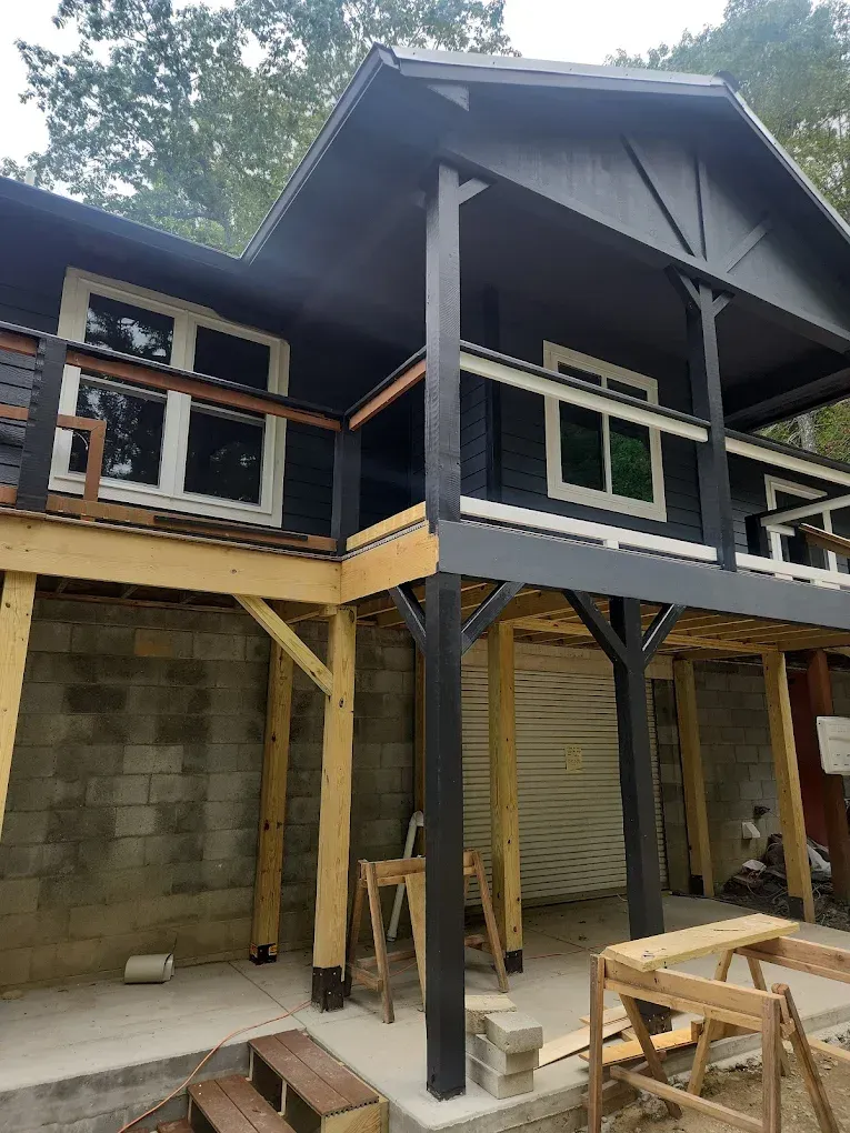 Partially built house with dark upper siding, exposed beams, and concrete lower walls under a covered porch