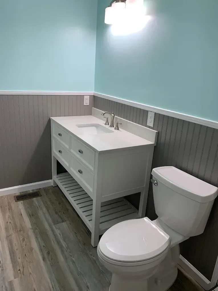 Small bathroom with white vanity, toilet, gray wainscoting, wood-look floor, and light blue walls.