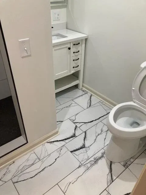 Small bathroom with white vanity, marble-pattern floor, and toilet beside an open shower or tub area.