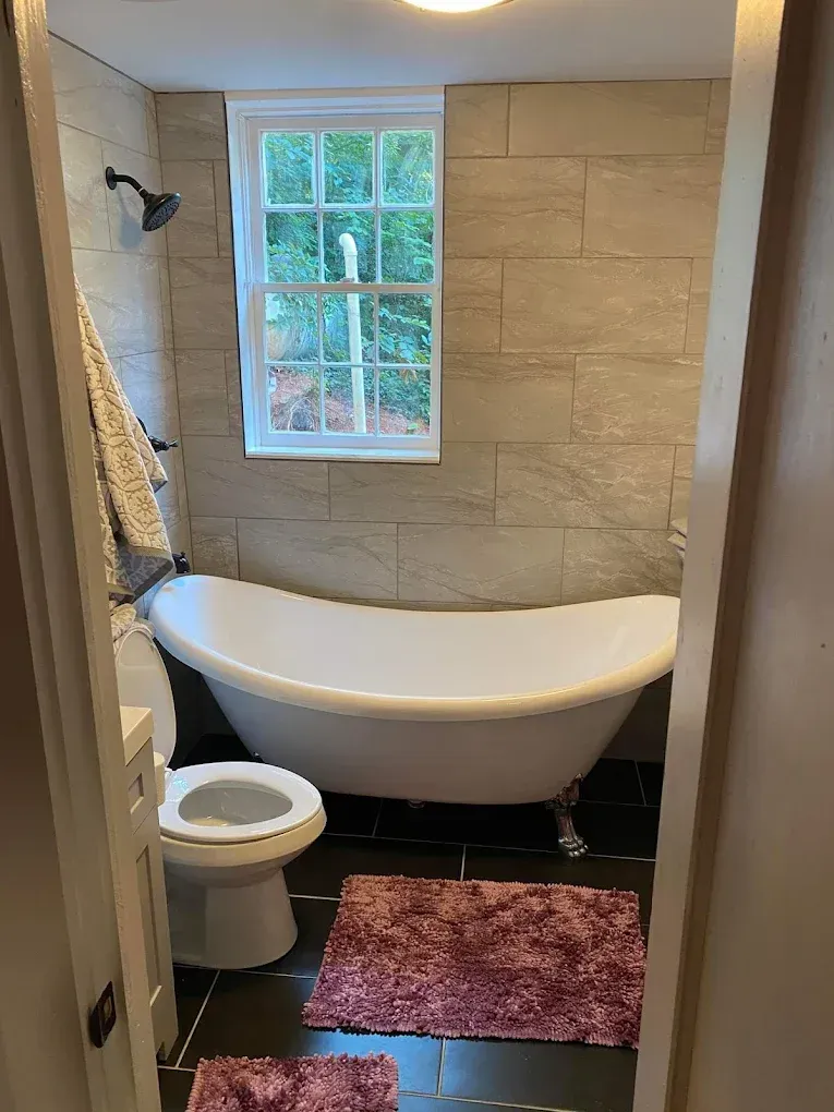 Bathroom with a white clawfoot tub, toilet, window, beige tile walls, and a red bath mat.
