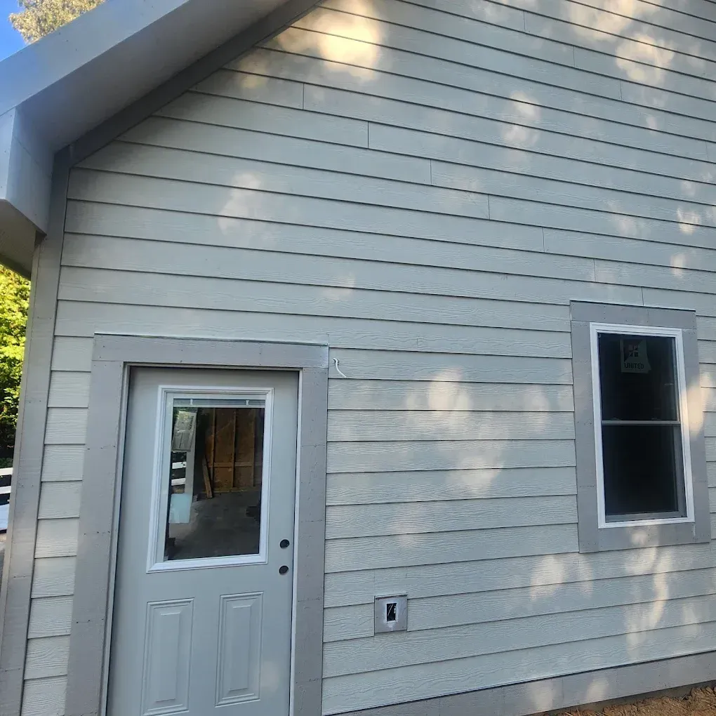 Small white house exterior with a side door and window, lit by sunlight and shadows from nearby trees