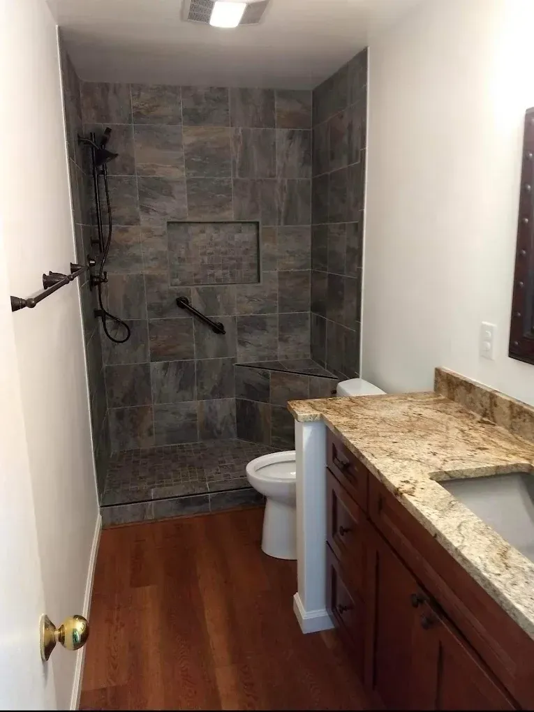 Narrow bathroom with tiled shower, white toilet, wood vanity, and granite countertop.