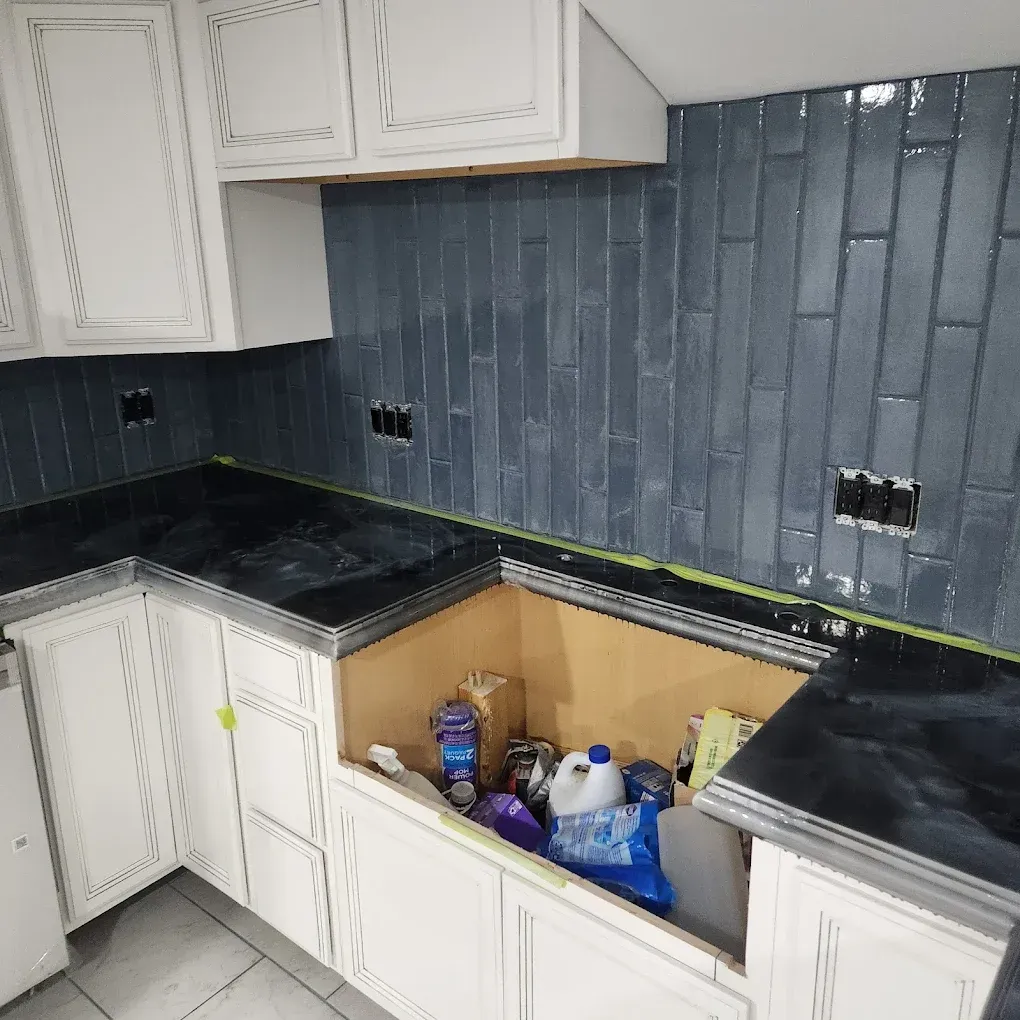 Kitchen with white cabinets, black countertops, and blue tile backsplash; lower cabinets are open and cluttered.