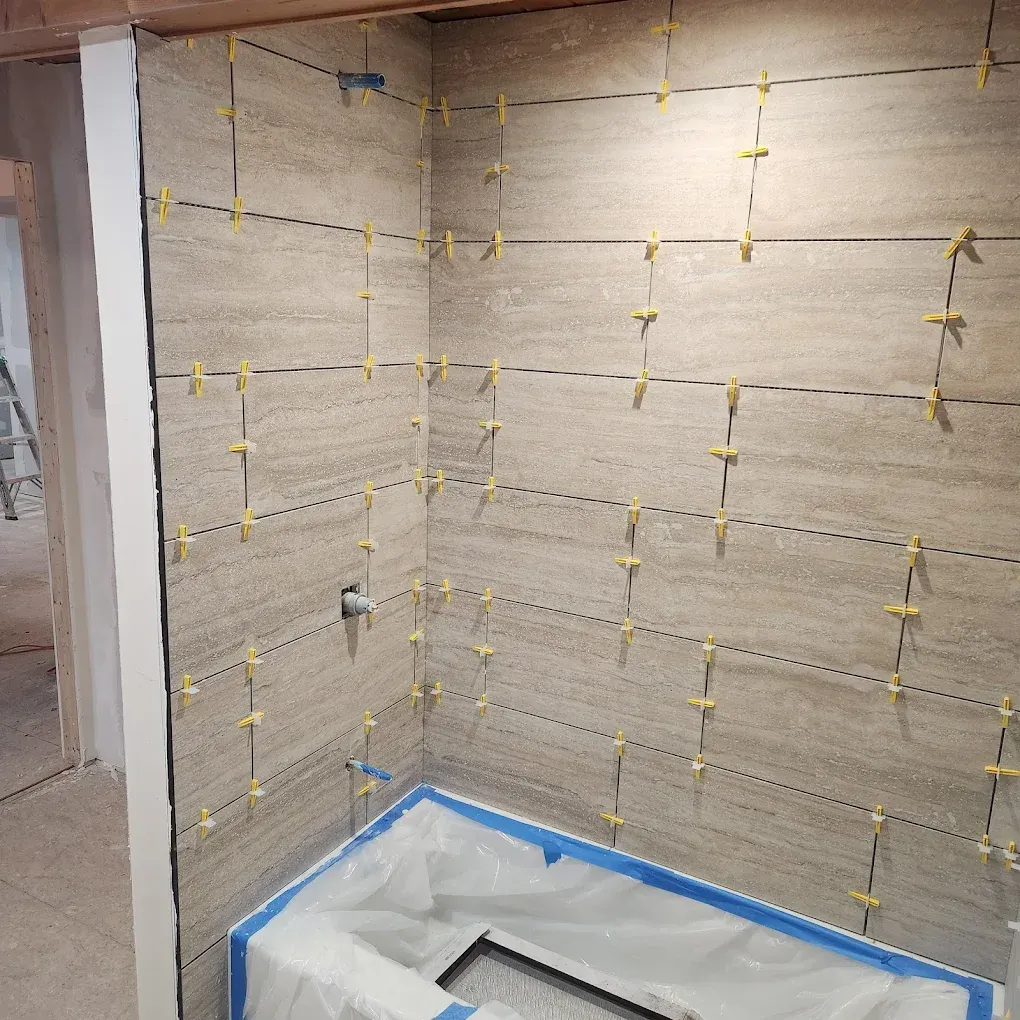 Partially tiled shower corner with beige stone-look tiles, yellow spacers, and blue painter’s tape at the base