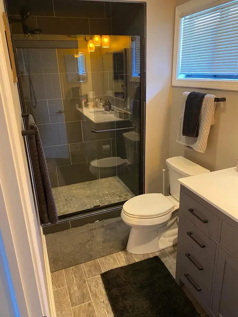 Bathroom with glass shower, toilet, vanity, and window with blinds.
