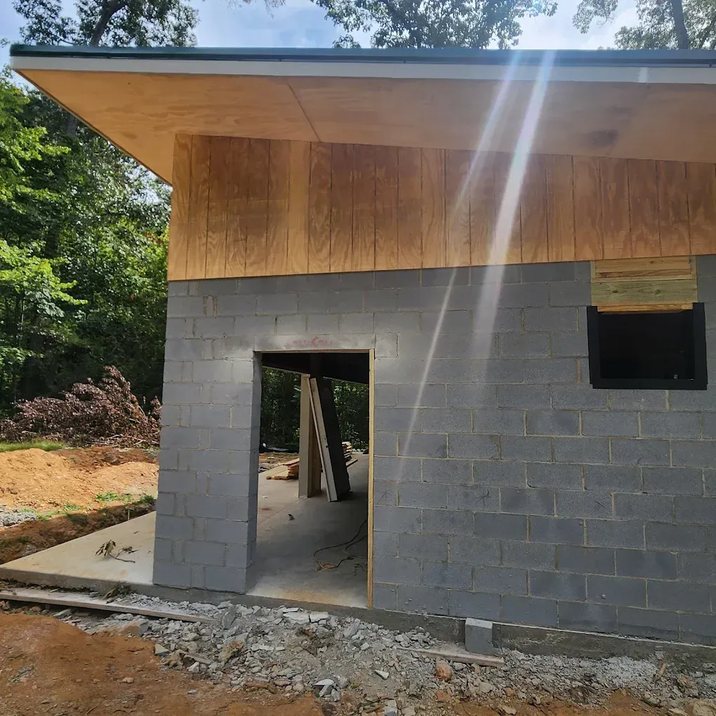 Partially built cinder block house with wood overhang, open doorway, and window in a sunny wooded area