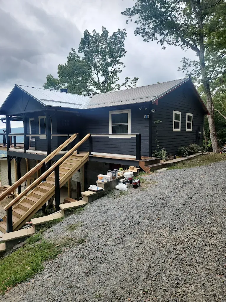 Black cabin with porch and wooden stairs on a gravel driveway, surrounded by trees under a cloudy sky