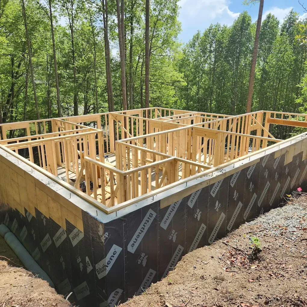 Wood-framed house foundation under construction on a black waterproofed basement wall in a wooded area