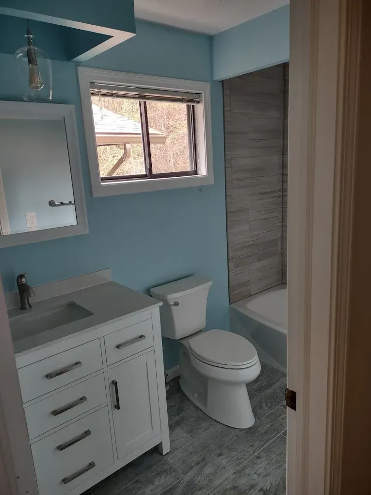 Small bathroom with light blue walls, white vanity, toilet, and tub with gray tile accent wall.