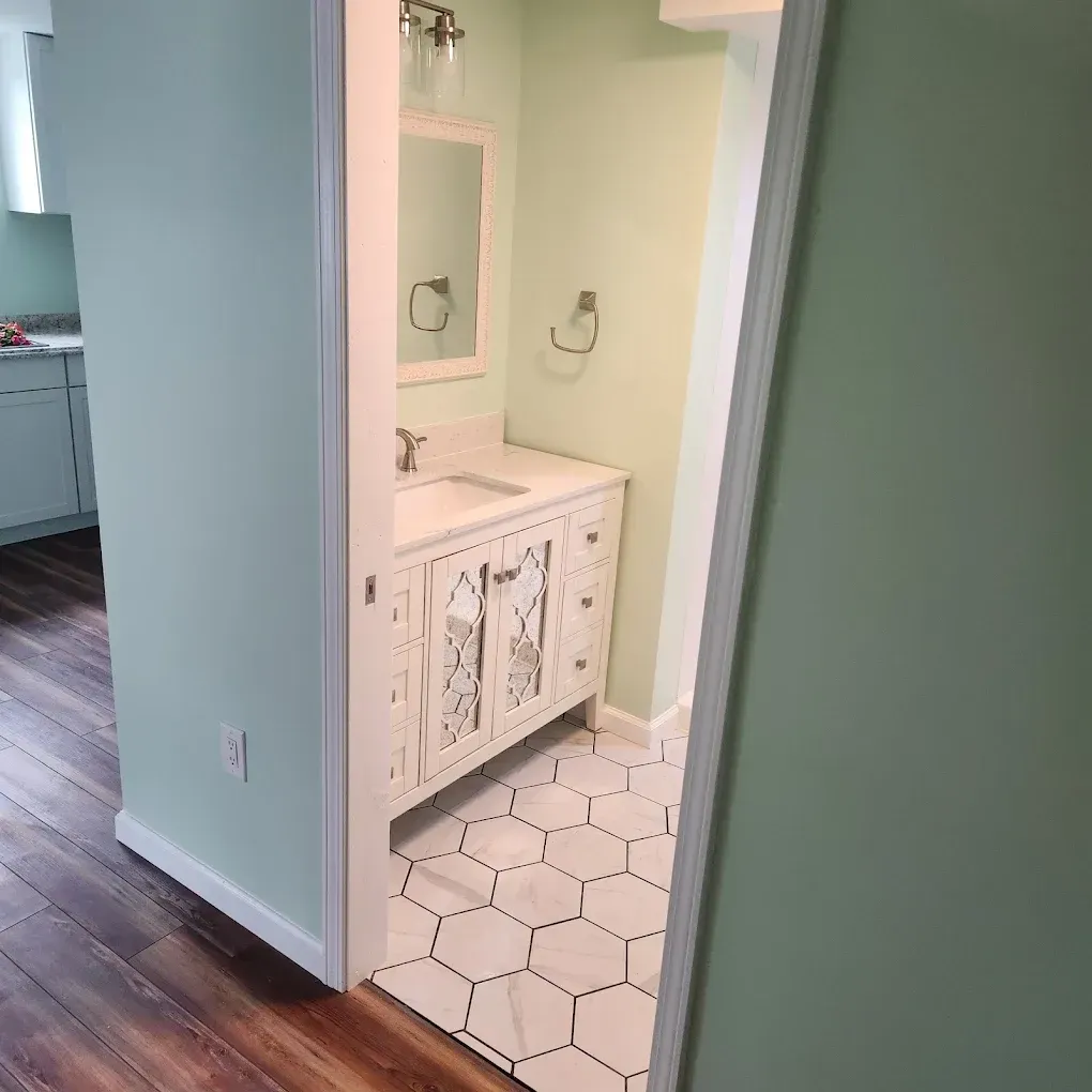Small bathroom vanity with white hex tile floor, visible through an open doorway from a wood-floored hall.