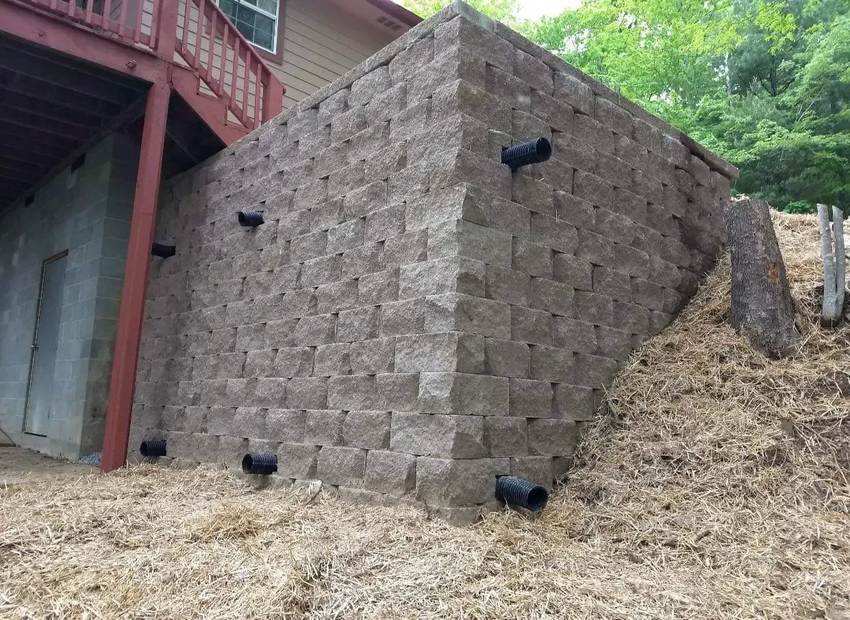 Stone retaining wall beside a red building on a slope, with leaf litter and trees nearby
