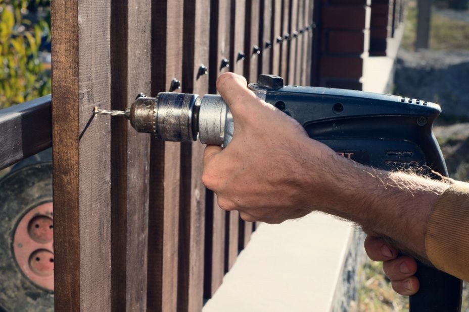A Man is Drilling a Hole in a Wooden Fence With a Drill — Mick's Carpentry & Maintenance In Machans Beach, QLD