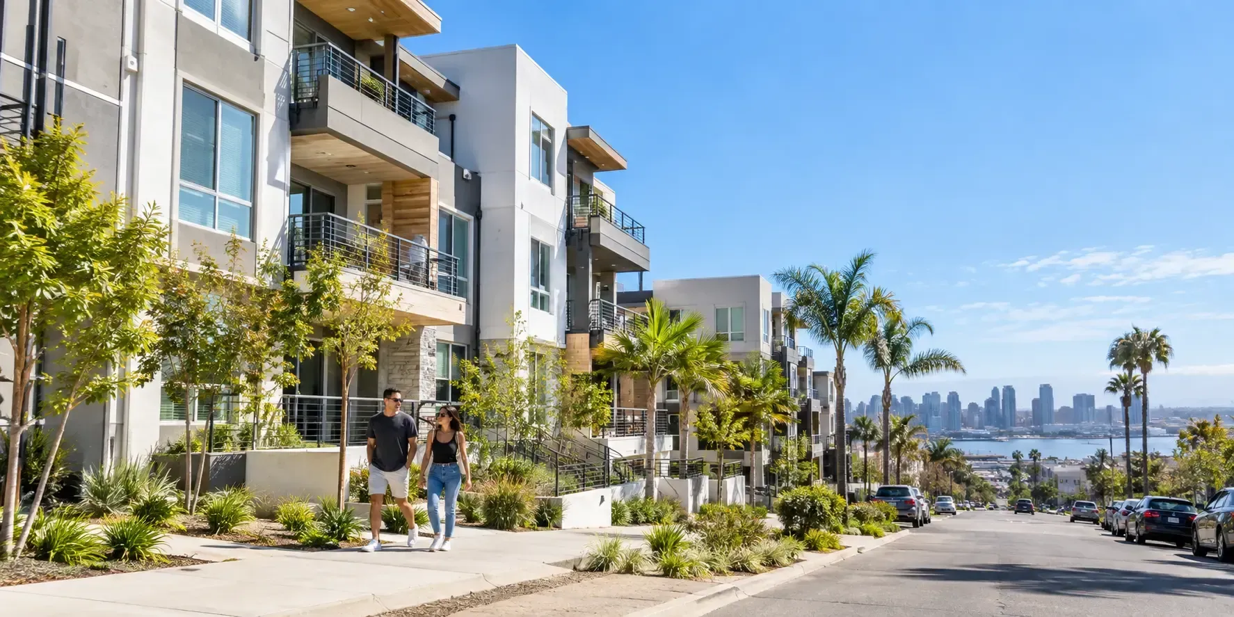 A modern multi-family apartment building in San Diego with balconies, landscaped walkways, palm trees, and residents walking under a clear blue sky.