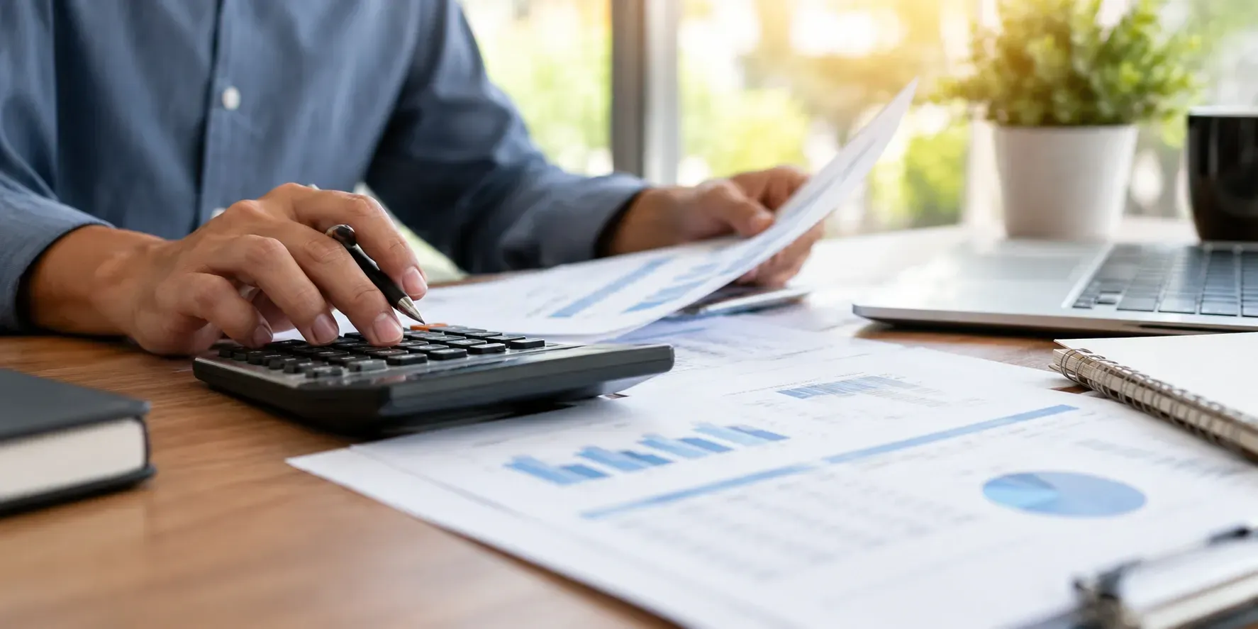 Close-up of a calculator, financial documents, and pen on a wooden desk with a professional reviewing paperwork in the background.