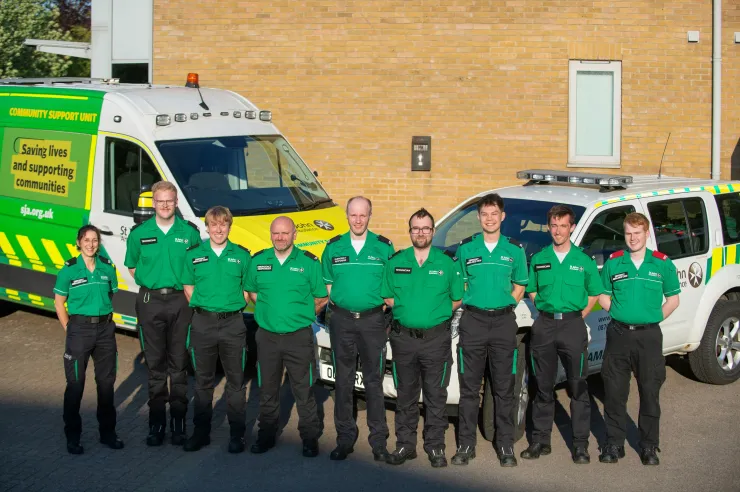 Nine people in green uniforms stand in front of ambulances outside a brick building.