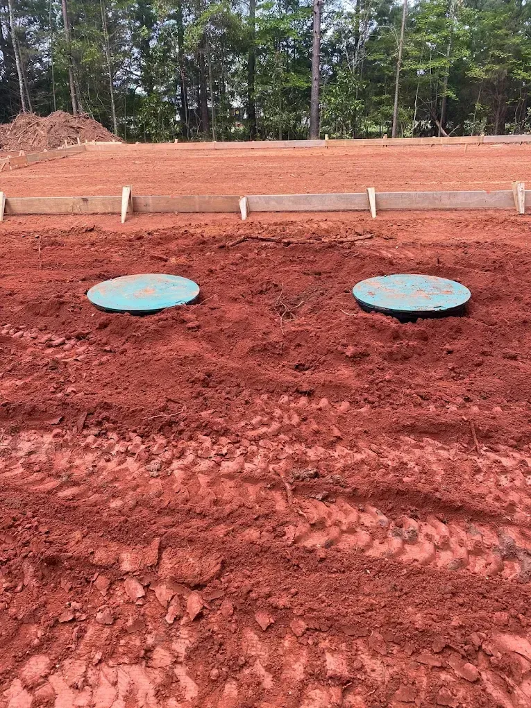 Two green circular covers in red dirt at a construction site, with wooden forms in the background.