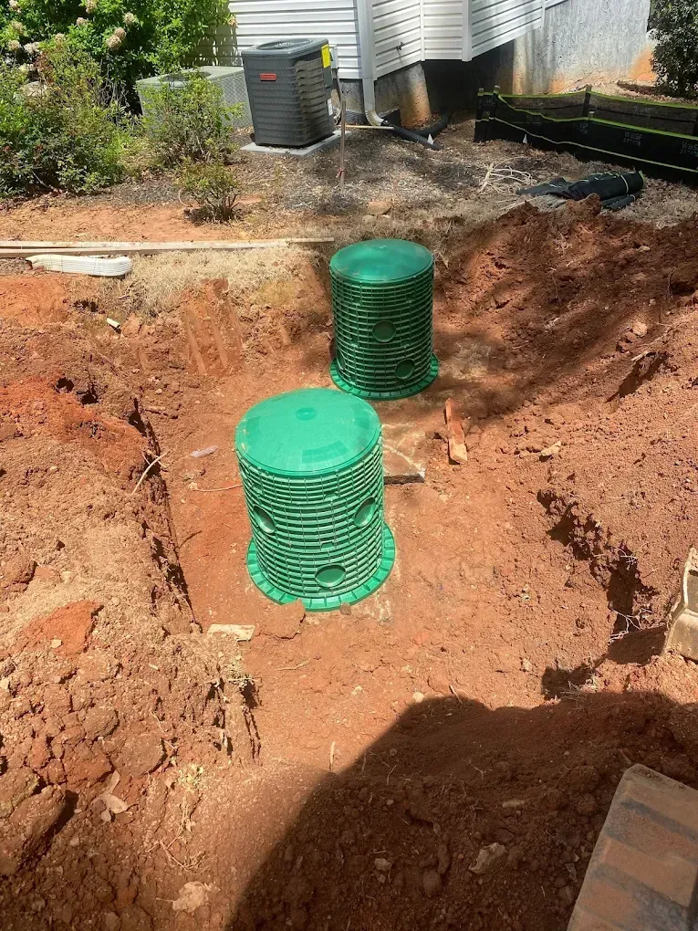 Two green septic tanks installed in a dirt excavation; a house and HVAC unit are in the background.