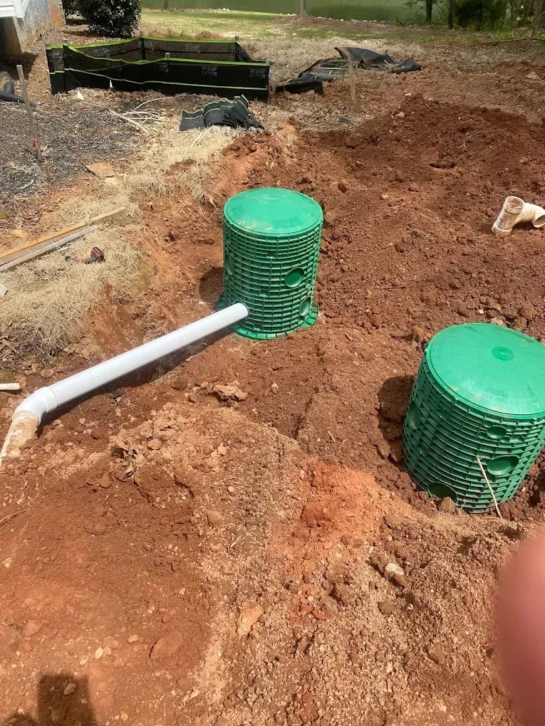 Two green plastic cylindrical structures in a dirt excavation with a white pipe leading into one.