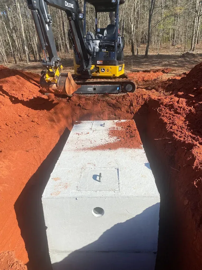 A small excavator places a concrete septic tank into a red-dirt trench.