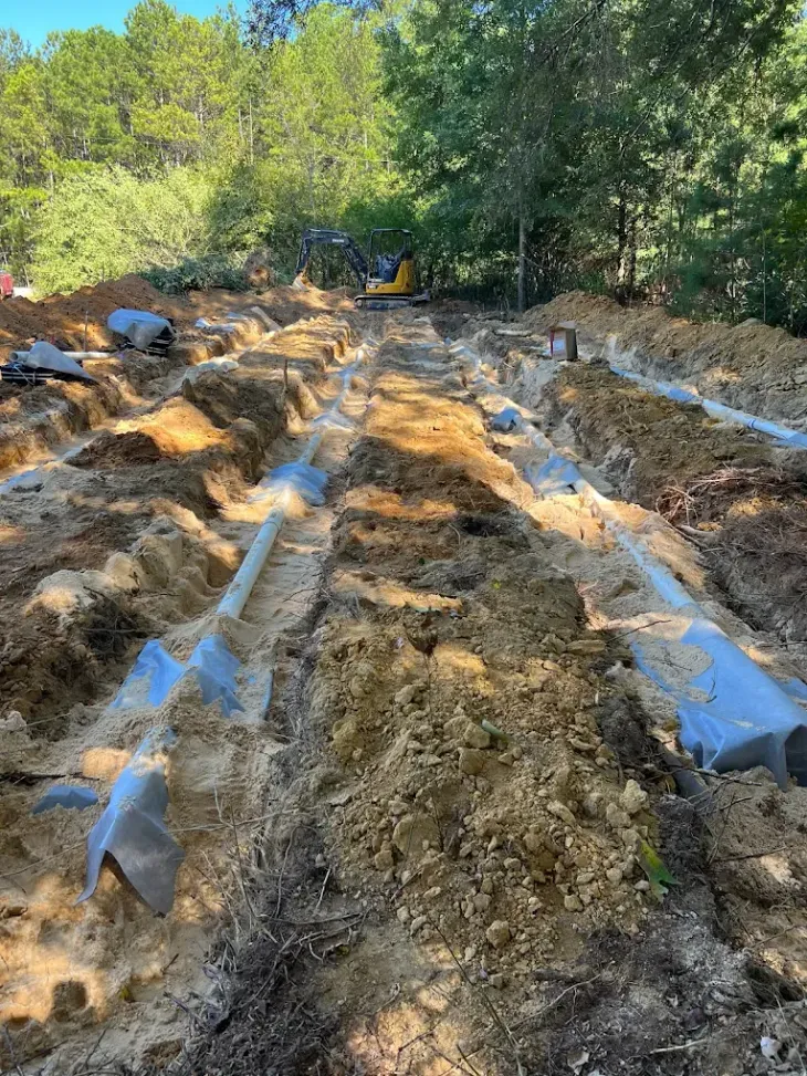 Trenches dug in soil, with gray pipes and a small excavator in a wooded area.