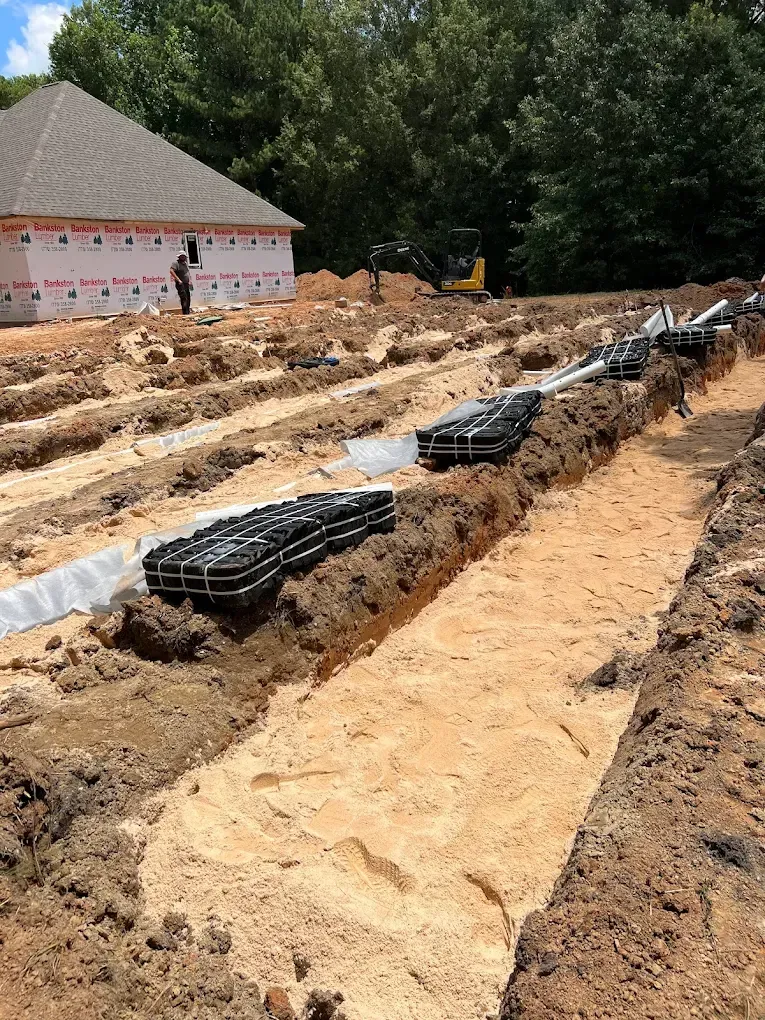 Construction site with trenches, drain fields, and new house under construction in background.