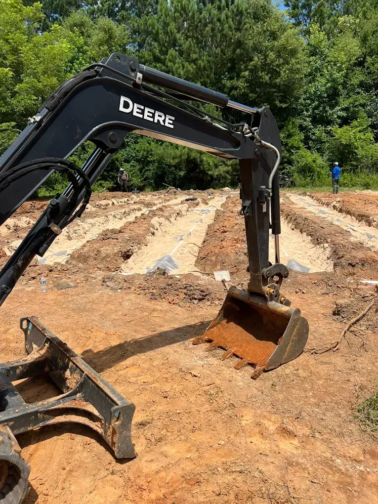 A John Deere excavator digging trenches in a dirt field. A person stands in the background.