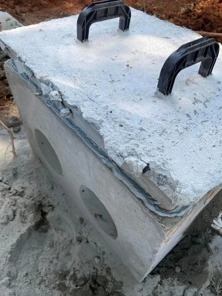 Concrete utility box with two circular openings and black handles on top, set in dirt.