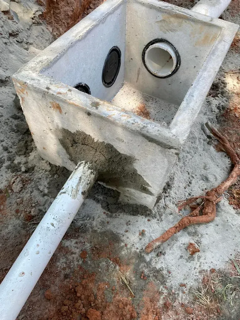 Concrete box with pipes, used for drainage, in dirt. A root lies nearby.