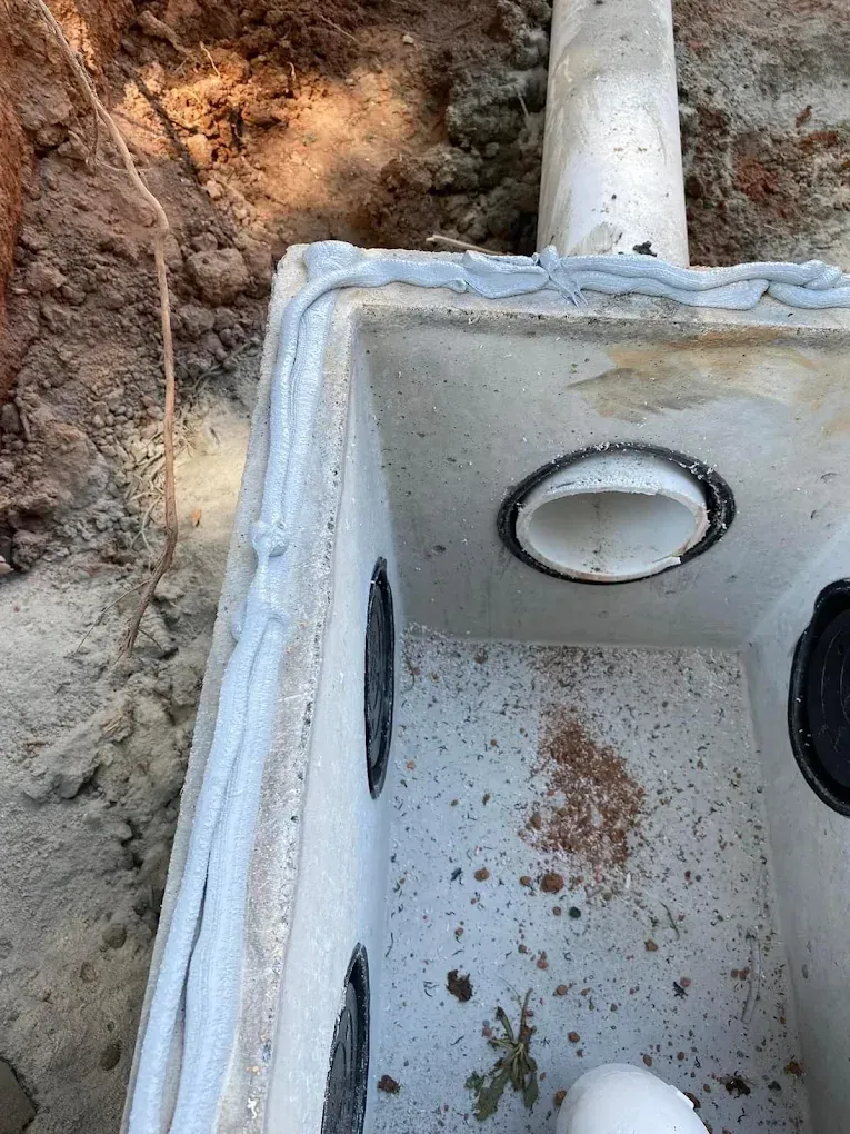 Concrete septic tank, open top, with PVC pipe and holes for connections, in a dirt pit.