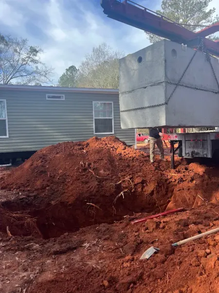 A concrete septic tank is lifted by a crane near a mobile home. A worker is near the excavated ground.