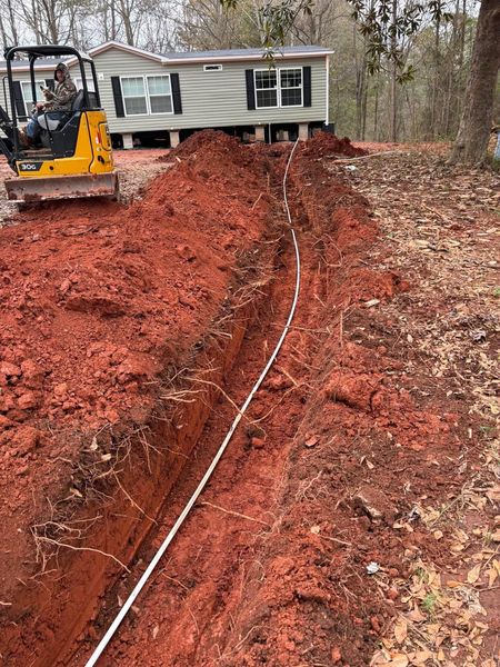 A trench dug in red soil, with white pipe laid in it. A yellow backhoe and a house are in the background.