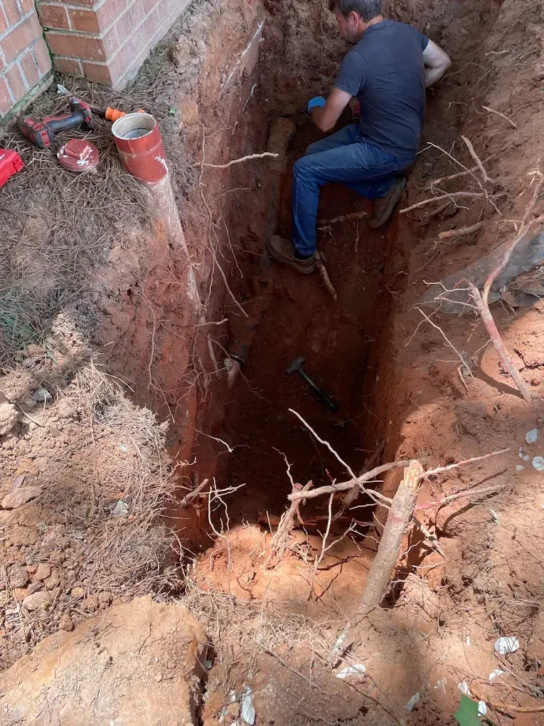 Man in blue shirt and jeans digging a deep trench next to a brick wall.