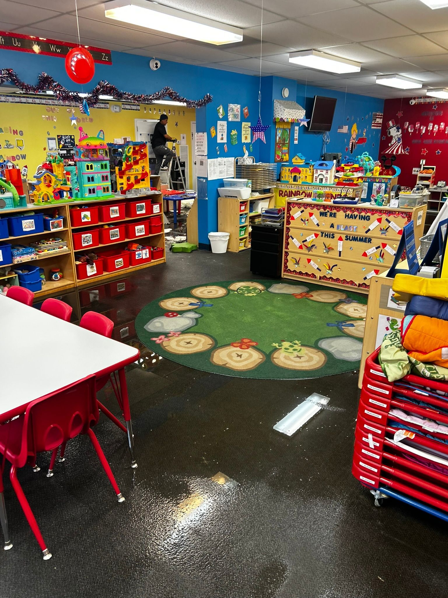 Colorful classroom with red tables, toy shelves, and a green rug in the center