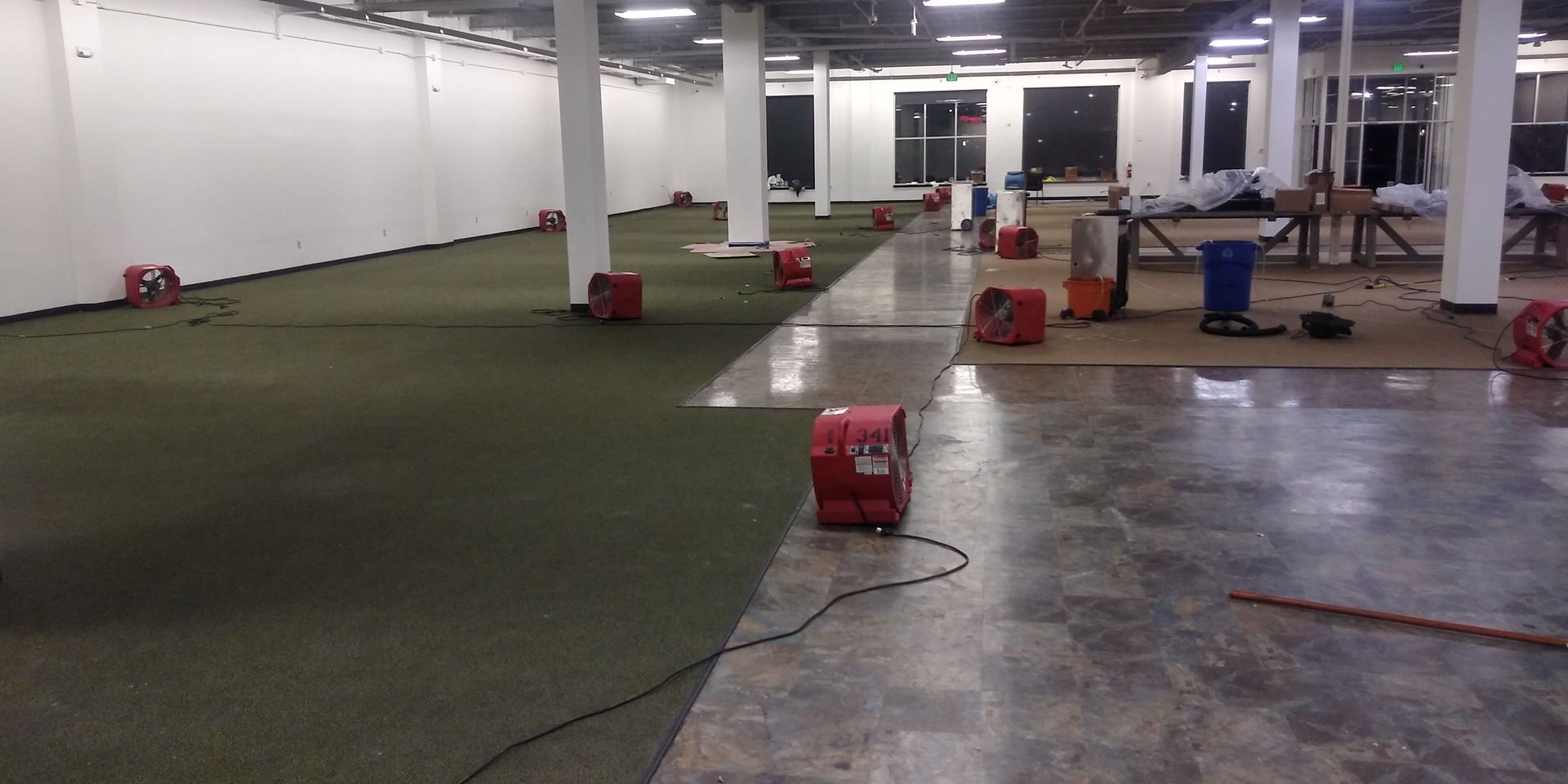 Large indoor room with green carpet, scattered equipment, and white walls under bright ceiling lights