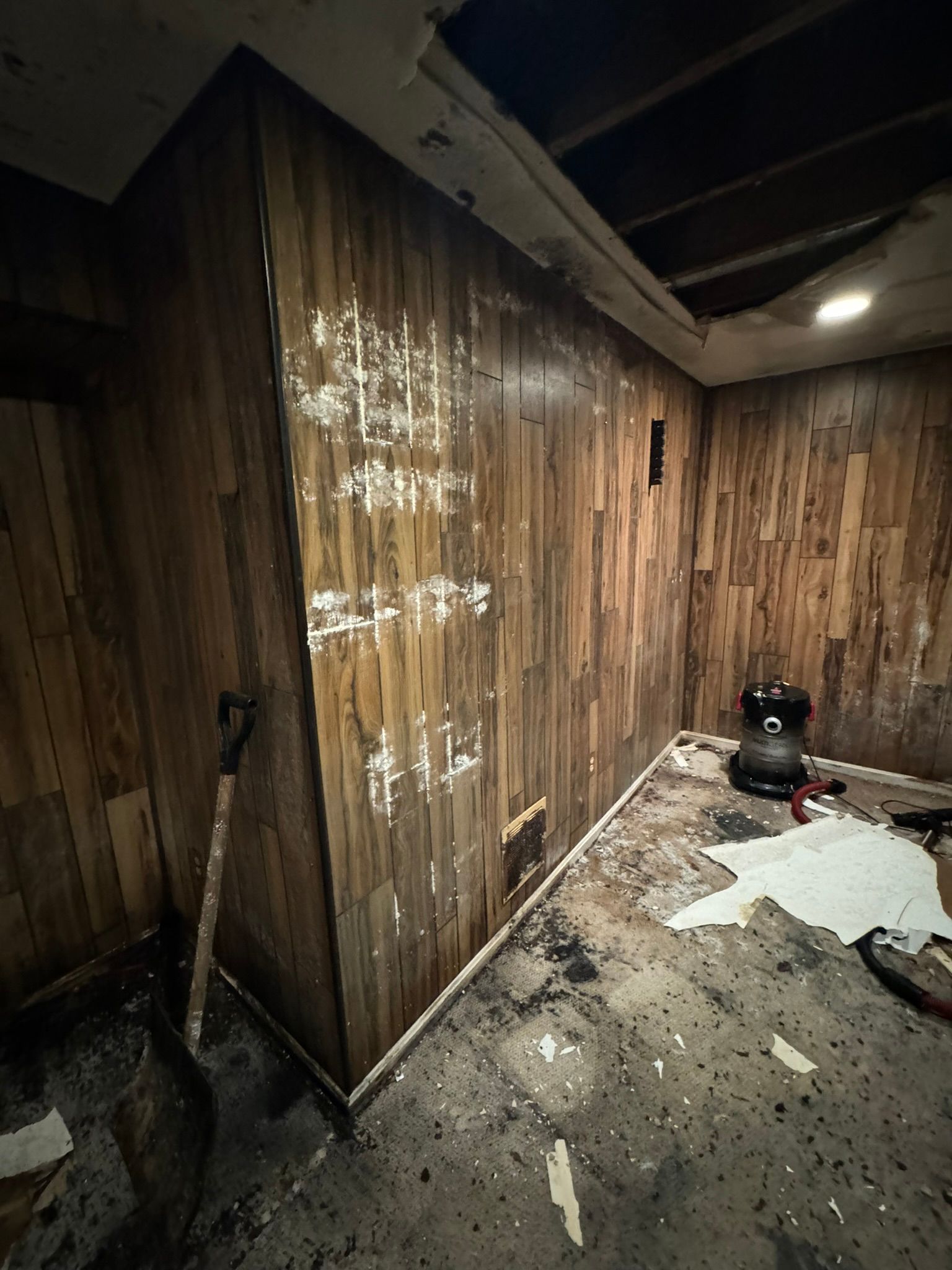 Damaged hallway with broken wall panels, scattered debris, and a mop leaning against the wall.