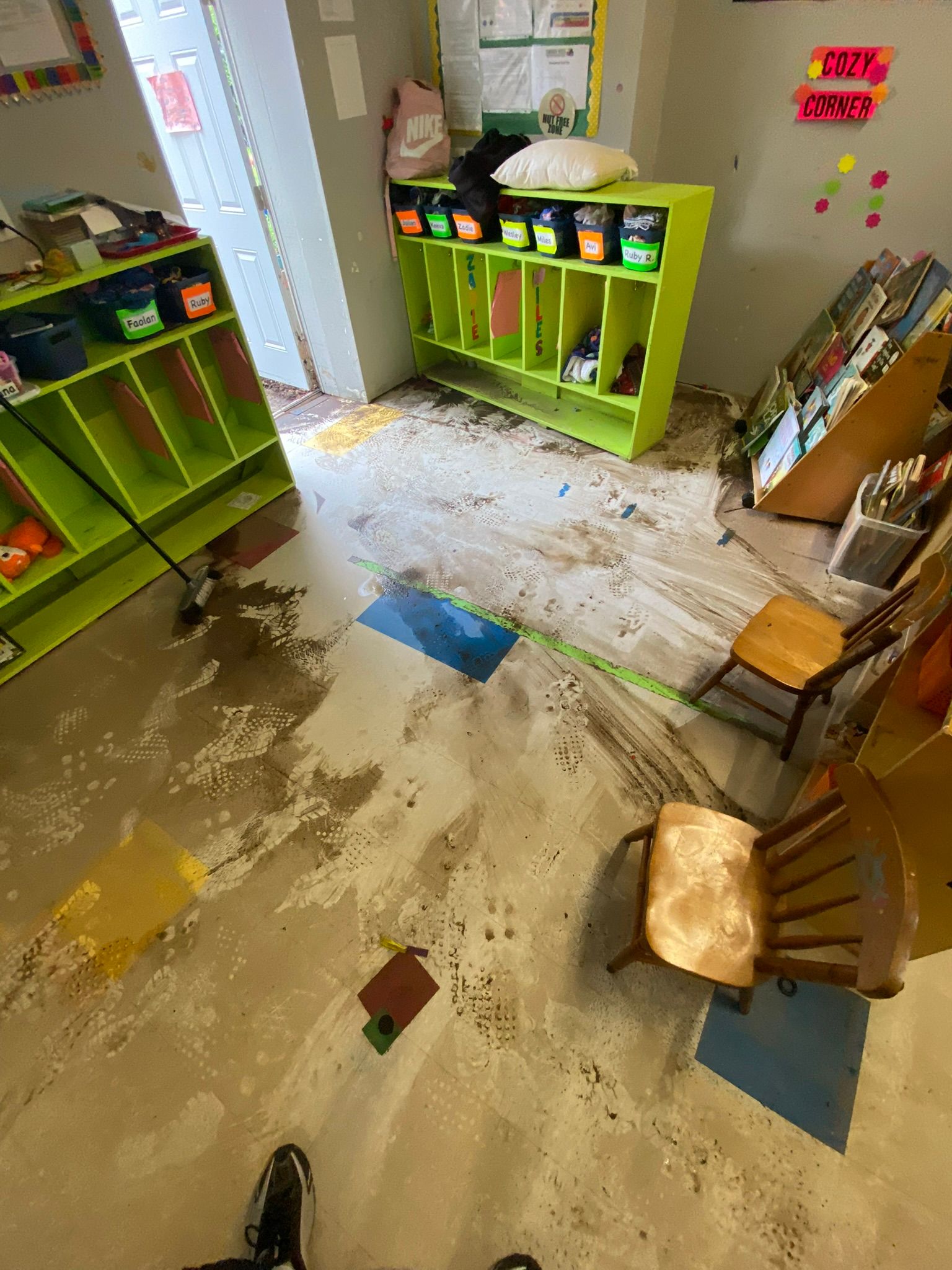 Messy classroom with scattered debris, tipped chairs, and green cubbies after a spill or damage