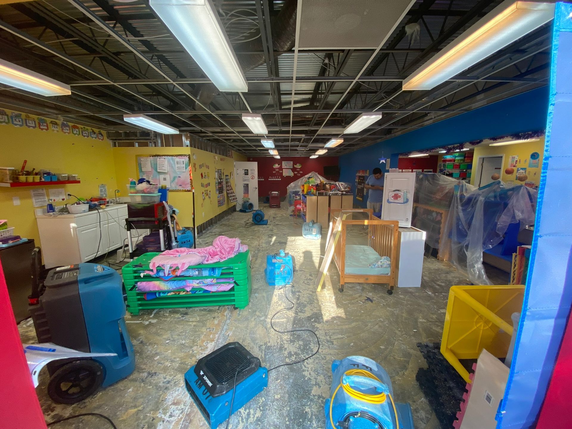 Indoor warehouse or play area with colorful bins, tables, and plastic-covered equipment under fluorescent lights