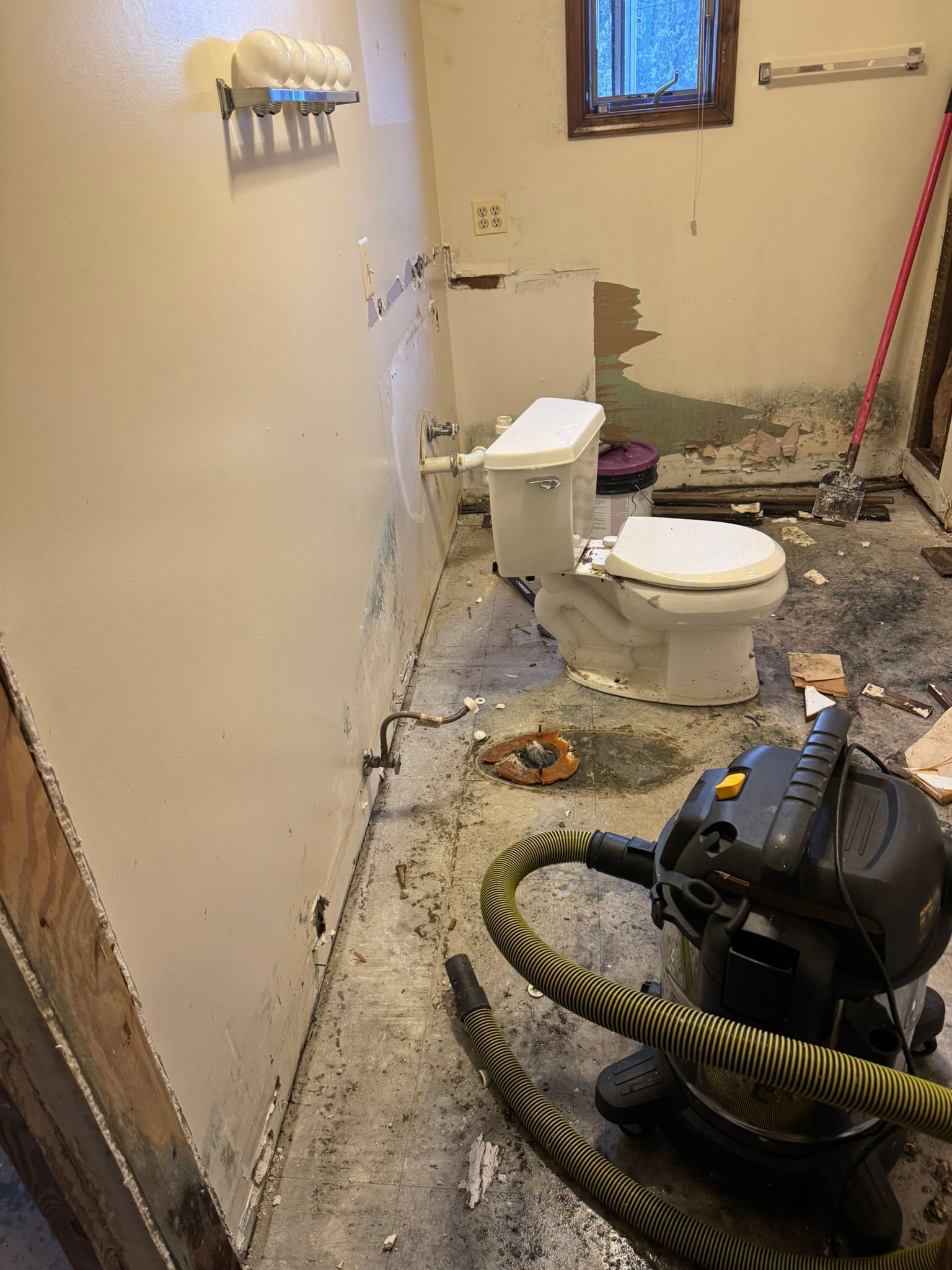 Damaged bathroom with a toilet, debris, and a wet vacuum on the floor, walls partially stripped