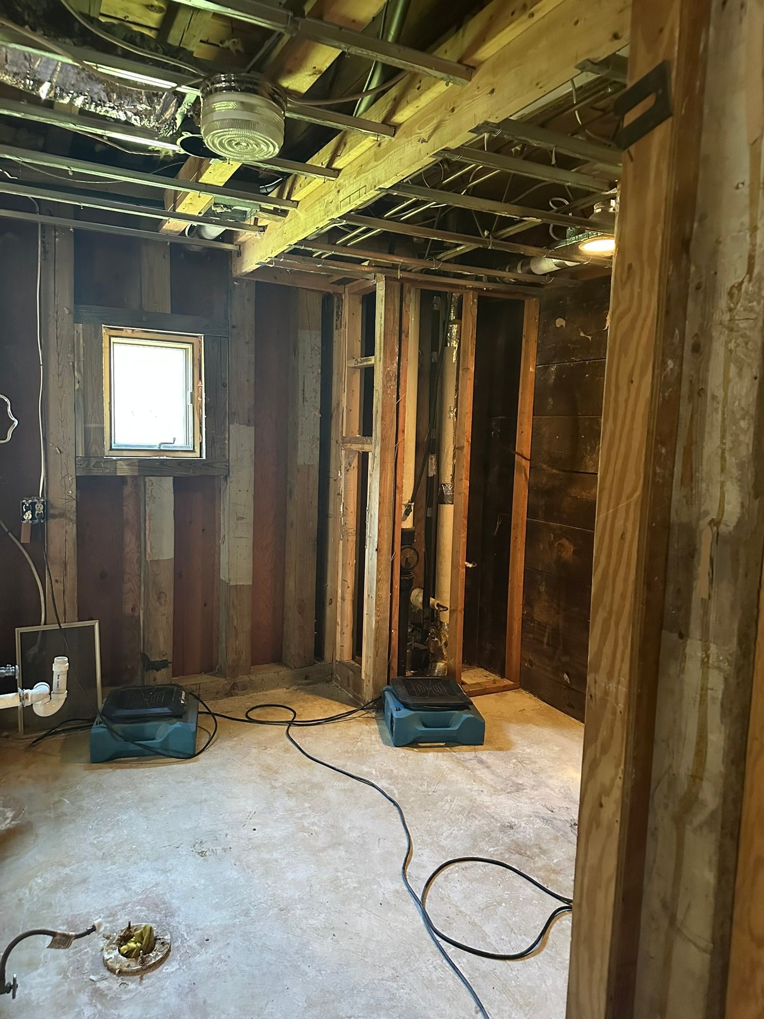 Damaged room with exposed wooden studs, broken ceiling, small window, and tools on a concrete floor