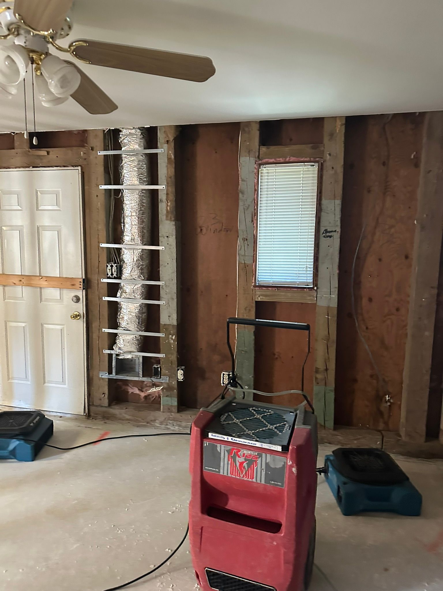 Partially stripped room with exposed studs, white door, window, fan, and red dehumidifier on the floor