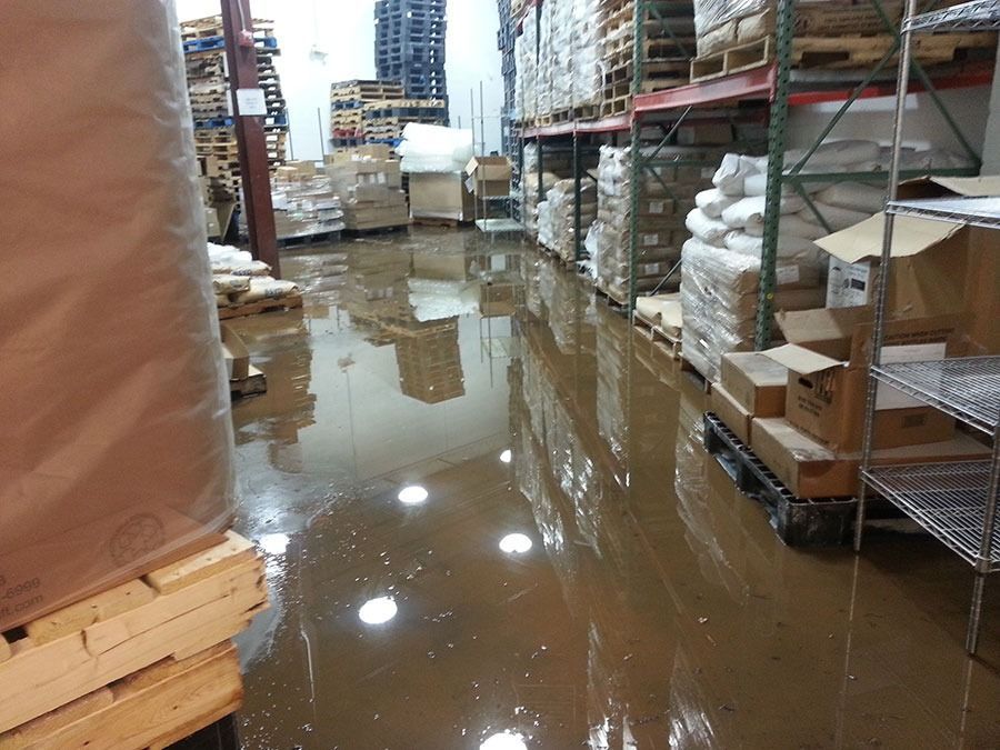 Flooded warehouse aisle with shelves, boxes, and standing brown water reflecting overhead lights